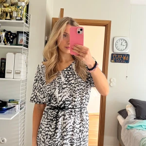 Svartvit mönstrad klänning med knytband - Snygg svartvit klänning med abstrakt mönster och korta ärmar. Den har ett knytband i midjan för en smickrande passform. Perfekt för en stilren look!