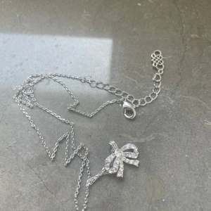 Lily and Rose Silverhalsband med rosett - Elegant silverhalsband med en glittrig rosett som hänge. Kedjan är tunn och justerbar med ett klassiskt lås. Perfekt för att lägga till en gnistrande detalj till din outfit.
