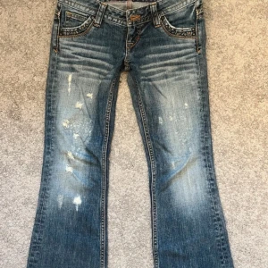 Blå bootcut jeans med slitningar - Snygga blå bootcut jeans med slitningar och dekorativa sömmar. Jeansen har en klassisk femficksdesign och är perfekta för en avslappnad stil. De har en låg midja och en lätt tvättad look.