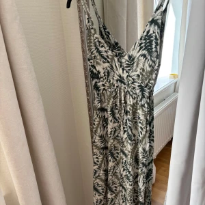 Långklänning med grönt bladmönster - Supersnygg långklänning med smala axelband och v-ringning. Klänningen har ett grönt och vitt bladmönster, knyt i midjan och är perfekt för sommardagar. Lätt och luftig känsla, passar dig som gillar en avslappnad men trendig stil. Okänt märke, köpt från en marknad i Belgien.