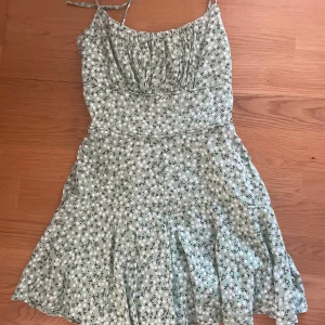 Grön blommig kortklänning med smala axelband - Supersöt ljusgrön kortklänning med små vita blommor och svarta detaljer. Klänningen har smala axelband och lätt rynkad byst, vilket ger en avslappnad och somrig vibe. Perfekt för varma dagar och picknickar i parken.