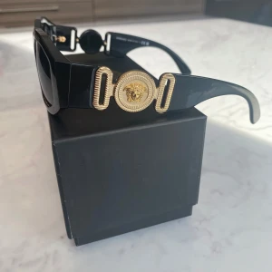 Svarta Versace solglasögon med guld - Snygga svarta solglasögon från Versace med rektangulära bågar och ikoniska guldiga Medusa-detaljer på sidorna. Glasögonen har en chunky design, perfekt för dig som vill sticka ut med en lyxig vibe.