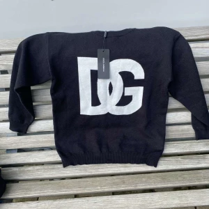Svart DG stickad tröja Dolce & Gabbana - Svart stickad tröja från Dolce & Gabbana med stor vit DG-logga framtill. Klassisk rund halsringning och långa ärmar. Perfekt för dig som vill ha en stilren men ändå iögonfallande look. Tröjan har ribbade muddar och är riktigt mjuk.