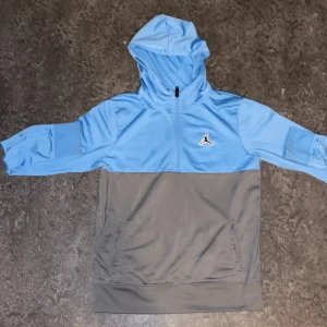 Jordan hoodie i blått och grått - Snygg hoodie från Jordan i ljusblått och grått. Tröjan har huva, half zip-dragkedja och den klassiska Jumpman-loggan på bröstet. Tillverkad i mjuk polyester, perfekt för chill eller sport. Fickor framtill och långärmad modell.