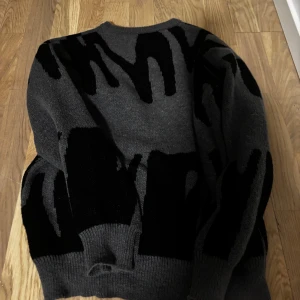 Grå stickad tröja med svart mönster - Säljer en grå stickad tröja med svart abstrakt mönster. Tröjan har rund halsringning och långa ärmar. 