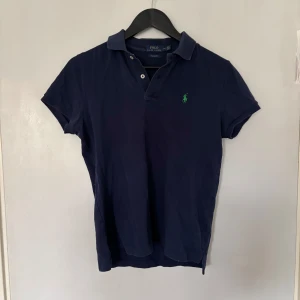 Marinblå pikétröja från Ralph Lauren - Klassisk marinblå pikétröja från Polo Ralph Lauren med korta ärmar och grönt broderat logotyp på bröstet. Tröjan har krage, två knappar och är tillverkad i bomull för en skön och avslappnad stil.