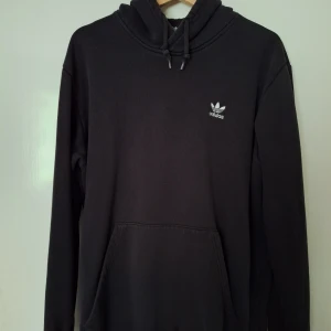 Svart Adidas hoodie - Svart hoodie från Adidas med klassisk vit trefoil-logga på bröstet. Hoodien har huva med snörning, känguruficka och långa ärmar. Mjuk insida och relaxed fit, perfekt för chill dagar. Har 2 små fettfläckar vid fickan som går att ta bort med en fläckborttagningspulver.