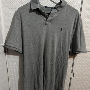 Grå pikétröja från Polo Ralph Lauren - Klassisk grå pikétröja från Polo Ralph Lauren med broderad grön logga på bröstet. Tröjan har korta ärmar, krage och knappar framtill. Tillverkad i mjuk bomull som är skön att bära. Perfekt för en clean och stilren look.