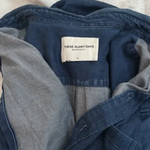 Jeans Skjorta från These Glory Days blå XL - Snygg mörkblå Jeans skjorta från These Glory Days i 100% bomull. Klassisk skjortkrage, bröstficka och knäppning framtill. Perfekt lager-på-lager-plagg med avslappnad vibe och stilren look.