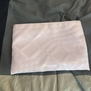 Rosa kuvertväska från Burberry - Säljer en stilren rosa kuvertväska från Burberry Fragrances. Väskan har ett diskret rutmönster och är tillverkad i glansig syntet. Stängs med dragkedja med metalltag. Perfekt att ha med sig när du vill hålla det enkelt men ändå classy.