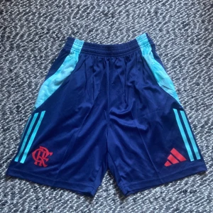 Flamengo shorts - HELT NYA Snygga marinblå sportshorts från Adidas med Flamengo-logga i rött och klassiska turkosa ränder på sidorna. Shortsen har elastisk midja och är gjorda i ett lätt, syntetiskt material som passar perfekt för fotboll eller träning.