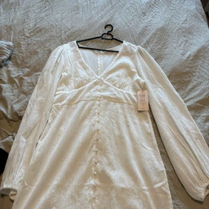 Vit långklänning från Nelly - Superfin vit långklänning från Nelly med v-ringning, långa ärmar och knappar framtill. Klänningen har en lätt struktur i tyget och känns både luftig och elegant. Perfekt för sommarens alla tillfällen!