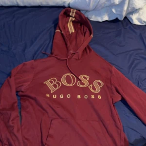 Vinröd hoodie från Hugo Boss - Snygg vinröd hoodie från Hugo Boss med stor gul logga framtill. Tröjan har huva med snörning och en klassisk känguruficka. Perfekt för dig som gillar streetstyle och vill sticka ut med ett exklusivt märke.