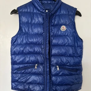 Blå dunväst från Moncler - Snygg blå dunväst från Moncler med quiltad design och hög krage. Västen har två dragkedjefickor framtill och klassisk Moncler-logga på bröstet. Perfekt att slänga över en hoodie för en cool look. Stängs med dragkedja hela vägen.