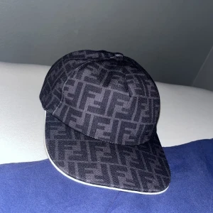 Svart Fendi keps med FF-mönster - Snygg svart keps från Fendi med det klassiska FF-logomönstret över hela kepsen. Kepsen har böjd skärm med vit kant och justerbar rem baktill. Tillverkad i bomullsmix som känns både lyxig och bekväm. Perfekt för dig som vill sticka ut med designerstil.