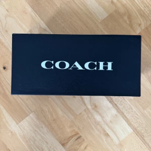 Rosa sandaler med klack från Coach - Snygga sandaler från Coach i en ljusrosa nyans med fyrkantig tå och låg klack. Skorna har en bred rem med en stor guldfärgad spänne som detalj. Tillverkade i skinn och perfekta för dig som vill ha en trendig och stilren look. Storlek 38/7.5 