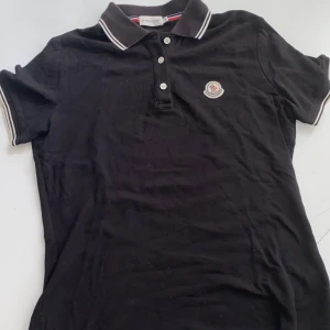 Svart Moncler pikétröja med logga - Svart pikétröja från Moncler med klassisk krage, vita ränder på krage och ärmslut samt broderad logga på bröstet. Tröjan har tre knappar framtill och är tillverkad i bomull för en skön och stilren look.