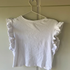 Vit ribbad topp med volangärmar - Superfin vit ribbad topp med volangdetaljer längs ärmarna. Toppen har en rund halsringning och är ärmlös med snygga volanger som ger en extra touch. 