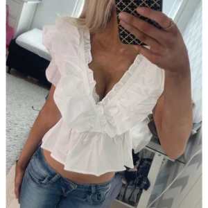 Vit volangblus från Meet Me There - Supersöt vit blus med djup v-ringning och stora volanger längs axlar och urringning. Modellen är croppad och ärmlös, perfekt för en trendig look. Tillverkad i lätt bomullstyg som känns fräscht och luftigt. Storlek xxs-xs aldrig änvandar