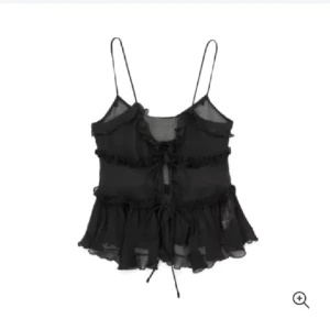 Svart volangig linnetopp från Zara - Supersöt svart linnetopp från Zara med tunna axelband, volangdetaljer och knyt framtill. Toppens tyg är lätt och transparent, vilket ger en luftig känsla. Perfekt för dig som gillar romantiska och trendiga plagg med extra detaljer.litet hål som inte syns när man har på plagget