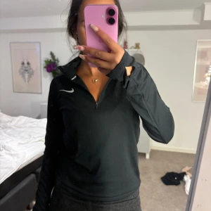 Svart Nike träningströja half zip - Svart långärmad träningströja från Nike med vit logga på bröstet. Tröjan har half zip-dragkedja och tumhål vid ärmsluten. Perfekt för löpning eller gym. Snygg och sportig passform i lätt syntetmaterial. Passar från Xs-M