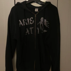 Svart hoodie med strass och dragkedja - Svart hoodie med huva och hel dragkedja framtill. Framsidan har ett coolt strassmönster med text och en siluett. Långa ärmar och känguruficka. Perfekt för dig som gillar streetstyle och vill sticka ut lite extra. Säljs pgd av att den är för liten.