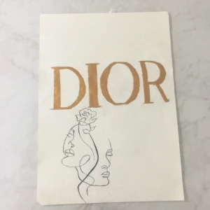 DIOR tavla  - Stilren bok med titeln 'DIOR' i guld och konstnärlig linjeteckning av två ansikten och en blomma. Perfekt för dig som gillar mode, konst och kreativitet. Passar som inspiration eller dekoration för unga vuxna och kreativa själar.