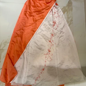 Lång axelbandslös klänning i satin - Snygg axelbandslös långklänning i orange och vitt satinliknande material. Klänningen har vita knappar i ryggen, broderade blommor och detaljer i vitt och orange. Perfekt för bal eller student. Sitter figurnära upptill och har vacker drapering.