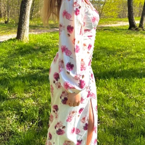 Blommig långklänning med slits - Superfin långklänning med blommigt mönster i rosa och rött på ljus botten. Klänningen har v-ringning, långa puffärmar, knappar framtill och en snygg slits. Perfekt för sommarens alla tillfällen!