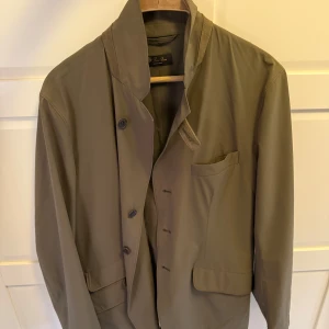 Loro Piana jacka - Snygg grön kavajjacka från Loro Piana med klassisk krage och flera fickor framtill. Jackan har knäppning med knappar och diskreta detaljer i mocka vid kragen. Insidan har en dragkedjeficka med röd accent. Perfekt för dig som gillar stilren och exklusiv look.