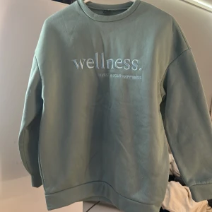 Grön sweatshirt med wellness-tryck - Mjuk grön sweatshirt med texten 'wellness. invest in your happiness' broderad framtill. Tröjan har rund halsringning och långa ärmar, perfekt för en chill och avslappnad stil.