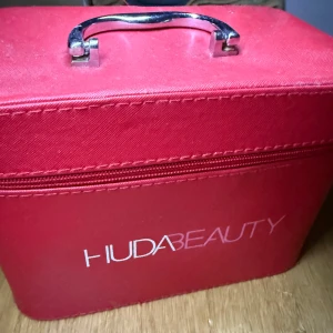 Röd sminkväska från Huda Beauty - Säljer en fyrkantig röd sminkväska från Huda Beauty med silvrigt handtag och dragkedja. Väskan är gjord i textil och har en rosa insida med plats för småsaker. Perfekt för att organisera smink eller accessoarer.