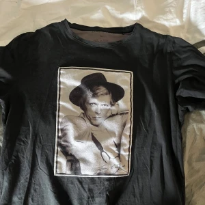 Limitato T-shirt - Svart t-shirt med ett stort fototryck på bröstet som föreställer en person med hatt. T-shirten har rund hals och korta ärmar. Trycket är i svartvitt och ger en cool vintagekänsla. Perfekt för dig som gillar unika motiv och streetstyle.