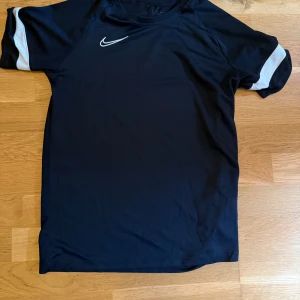 Svart Nike t-shirt med vita detaljer compression  - Svart t-shirt från Nike med klassisk vit swoosh-logga på bröstet och vita paneler på ärmarna. Sportig design i lätt och bekvämt material, perfekt för träning eller chill. Kortärmad modell med rund hals.