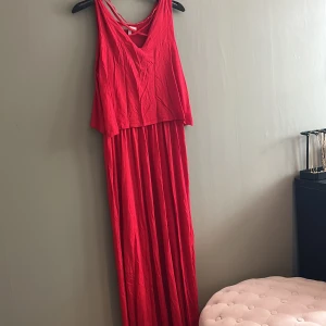 Röd långklänning från Sincerely Loved - Snygg röd långklänning från Sincerely Loved med smala axelband och v-ringning fram. Klänningen har ett lager på lager-utseende upptill och är ärmlös, perfekt för varma dagar. Tillverkad i mjukt och lätt tyg som faller fint.