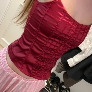 Vinröd satin korsettopp med smala axelband - Säljer en vinröd korsettopp i glansig satin med smala axelband. Toppen har markerade sömmar som ger en snygg formad look och sitter tight mot kroppen. Perfekt för dig som vill sticka ut med en elegant och trendig vibe.