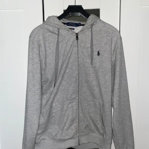 Grå hoodie från Polo Ralph Lauren - Snygg grå hoodie från Polo Ralph Lauren med dragkedja, huva och snörning. Klassisk broderad logga på bröstet och två fickor framtill. Custom fit-modell i mjuk bomull, perfekt för chill dagar eller när du vill ha en clean look.