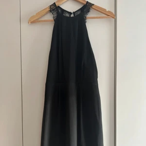 Svart klänning med spetsdetaljer - Supersnygg svart klänning med spets vid axlarna och öppen rygg. Klänningen har en rak, något lös passform och är ärmlös. Perfekt för dig som vill ha en stilren look med en touch av elegans