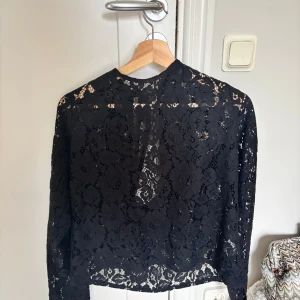 Svart spetsblus från Bik Bok - Supersnygg svart spetsblus från Bik Bok med lång ärm och blommigt spetsmönster. Blusen har öppen rygg med knytband i nacken och en rak passform. Perfekt för dig som vill ha en cool och elegant look med extra detaljer.