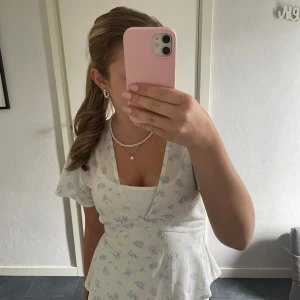 Blommig omlottblus från Gina Tricot - Supersöt vit blus med ljusblått blommönster från Gina Tricot. Modellen har v-ringning, korta ärmar och knytband i midjan för en snygg passform. Perfekt för vår och sommar, lätt och luftig i bomullsmix.