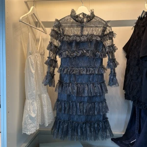 By Malena  - Supersnygg mörkgrå kortklänning i spets med flera lager volanger och hög krage. Klänningen har långärmade ärmar i transparent spets och en figurnära passform. Perfekt för dig som vill sticka ut med en unik och trendig look. Endast använd en gång. Köpt för 2800kr