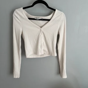 Vit croppad ribbad topp från NA-KD - Säljer en vit croppad topp från NA-KD med lång ärm och v-ringning. Toppen är ribbad och har knappar framtill för en snygg detalj. Perfekt att styla med jeans eller kjol för en trendig look.