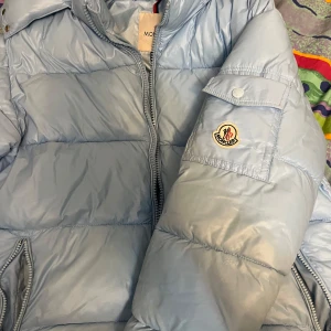 Ljusblå pufferjacka från Moncler - Säljer en snygg ljusblå pufferjacka från Moncler med klassisk logga på ärmen. Jackan har dragkedja framtill, hög krage och praktiska fickor. Perfekt för kalla dagar och riktigt skön att bära. Fodret har en cool serietidningsdetalj på insidan.