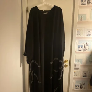 Svart långklänning med silverdetaljer - En svart långklänning med vida ärmar och rund halsringning. Klänningen har snygga silverfärgade broderade detaljer längs nederdelen och ärmarna som ger en cool touch. Passformen är loose och tyget faller mjukt. Perfekt för dig som gillar en stilren och bekväm look.