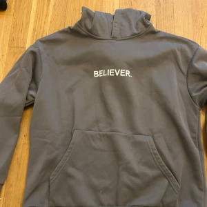  hoodie med texten BELIEVER. - Snygg grå hoodie med trycket 'BELIEVER.' på bröstet. Tröjan har en stor känguruficka framtill och hög krage. Perfekt för dig som gillar en enkel men cool stil.