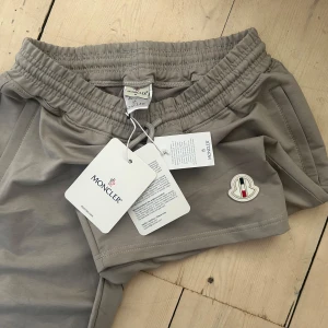 Grå Moncler shorts med snörning - Snygga grå shorts från Moncler med elastisk midja och snörning. Klassisk design med fickor på sidorna och bak, samt Moncler-logga framtill. Perfekta för sommaren och chill dagar. Materialet känns mjukt och lätt, riktigt sköna att bära.