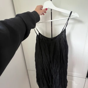 Svart omlottklänning med tunna axelband - Supersnygg svart kortklänning från Sisters Point med tunna axelband och lätt skrynklig look. Klänningen har omlottkänsla och är perfekt för dig som gillar en enkel men trendig stil. Materialet känns lätt och flowy.