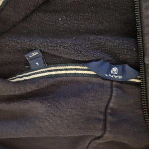 Marinblå hoodie från GANT med dragkedja - Snygg marinblå hoodie från GANT med vit logga på bröstet. Tröjan har huva med vita snören, hel dragkedja och två fickor framtill. Perfekt för en chill och sportig look. Tillverkad i mjuk bomullsmix för extra komfort. Lite urtvättad men inget som märks mycket.