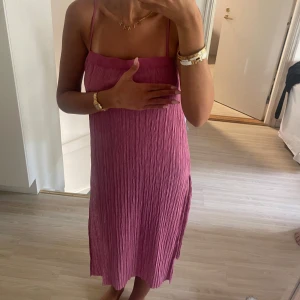 Plisserad rosa klänning med smala axelband - Säljer en plisserad rosa klänning från zara med smala axelband och rak halsringning. Klänningen är midi-lång och har en lätt glansig yta som ger en lyxig känsla. Perfekt för sommarens alla tillfällen och enkel att styla med smycken. Har en slits nertill 