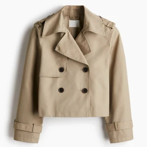 Beige croppad trenchcoat med knappar - Stilren croppad trenchcoat i beige med dubbelknäppning och bred krage. Jackan har långa ärmar med spännen vid ärmsluten och axelklaffar för en klassisk look. Perfekt för dig som vill ha en trendig och tidlös jacka.